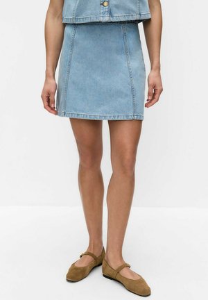 Kvinde iført lysblå denim mininederdel og matchende cropped jakke med brune ruskind Mary Jane-flats på hvid baggrund.