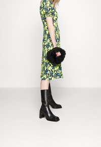 Persona in un vestito floreale giallo che tiene una borsa nera pelosa, indossando stivali di pelle con tacco fino al polpaccio neri, in piedi su un pavimento grigio.