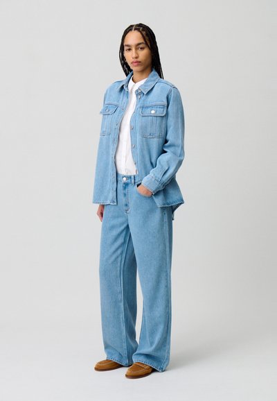 Giacca di jeans blu chiaro con due tasche sul petto, abbinata a jeans a gamba larga coordinati e scarpe marroni. Semplice camicia bianca sotto.