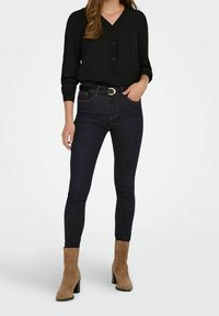 Camicia nera a bottoni abbinata a jeans skinny blu scuro e stivaletti alla caviglia in suede beige con tacco a blocco. Design semplice e aderente.