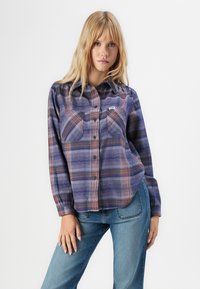Young woman wearing purple and orange plaid button-up shirt with long sleeves and blue jeans, standing against a plain white background.