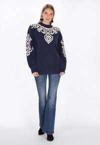 Marineblauwe trui met een crèmekleurig decoratief patroon, inclusief harten, ronde hals en lange mouwen. Gecombineerd met uitlopende blauwe jeans en zwarte laarzen.