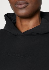 Felpa nera con cappuccio in tessuto testurizzato, un cappuccio spazioso e il marchio "Calvin Klein" impresso sul davanti. Vista ravvicinata che evidenzia i dettagli di design.