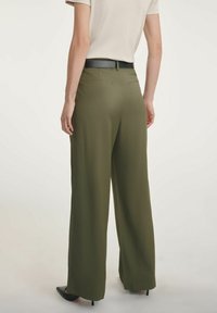 Oliwkowe spodnie o szerokich nogawkach z gładką teksturą, z dwoma tylnymi kieszeniami, w połączeniu z czarnym paskiem i czarnymi szpilkami.