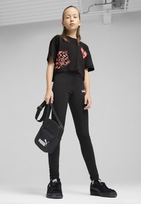 Černý crop top s růžovým leopardím vzorem, černé elastické legíny a černé tenisky, držící malou černou kabelku přes rameno s logem Puma.