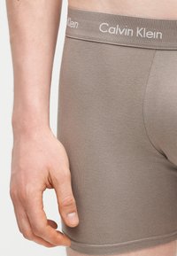 Κοντινό πλάνο ενός χεριού που ακουμπά δίπλα σε μπεζ μποξέρ Calvin Klein με λογότυπο στη μέση.