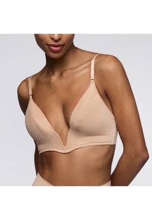 Femme portant un soutien-gorge plongeant beige avec des bretelles réglables, montrée des épaules au ventre sur un fond clair uni.