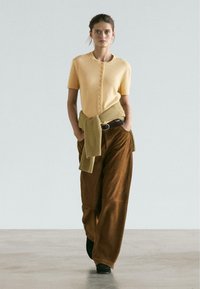Yellow knitted short-sleeve sweater with button detailing, paired with brown wide-leg pants. Sweater tied at the waist with ribbed texture.