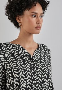 Blusa nera con un motivo astratto bianco, design a V, e una fila di bottoni. Il tessuto ha una trama liscia con una leggera lucentezza.