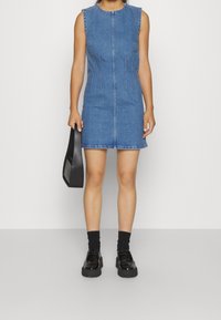 Vestido recto de mezclilla con silueta ajustada, escote redondo, diseño sin mangas y detalles de costura visibles. Combinado con zapatos de plataforma negros y bolso.