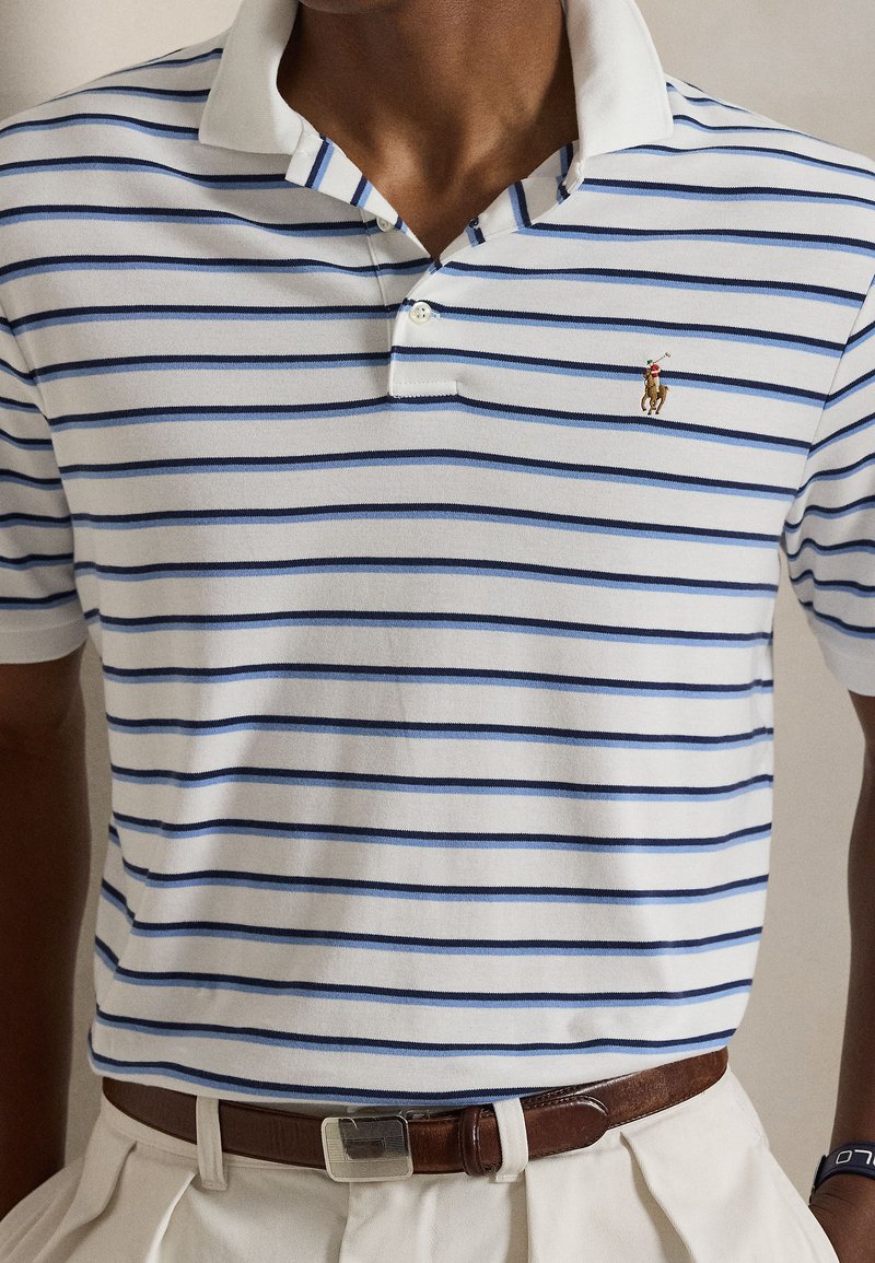 Polo à rayures blanches et bleu marine, manches courtes, col côtelé, et petit logo de cheval brodé sur la poitrine. Pantalon beige avec ceinture.