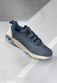 Chaussure de sport bleue avec surface texturée, design incurvé, lacets noirs et accents beiges. La semelle en caoutchouc présente un motif de camouflage subtil.