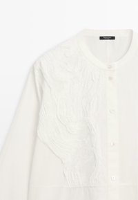 Cardigan blanc boutonné avec encolure ronde et motif subtil en vague texturée sur l'épaule gauche et la poitrine.