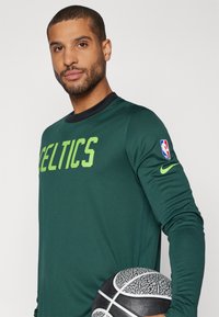 Žalias ilgas rankoves NBA marškinėliai su žodžiu "CELTICS" ryškiomis žaliomis raidėmis, juoduoju apykakle ir Nike logotipu ant rankovės, laikomi šalia krepšinio.