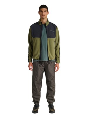 Jonge man met een olijfgroene en zwarte jas met ritssluiting, groen t-shirt, donkerbruine cargobroek en groen-zwarte sneakers, staande met het gezicht naar voren.