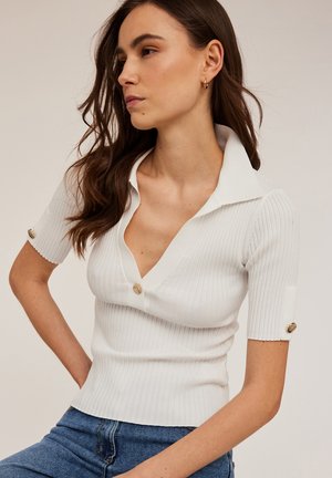 Top bianco a coste con scollatura a V profonda, maniche corte e bottoni dorati decorativi ai polsini. Il design aderente mette in risalto la silhouette.