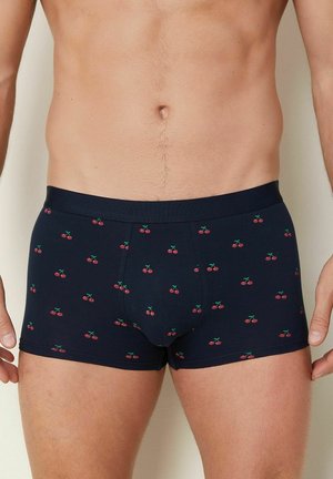 Torso maschile e cosce superiori che indossano boxer blu navy con un motivo di ciliegie rosse, in piedi contro uno sfondo chiaro e semplice.