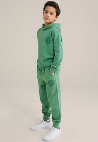 Conjunto de sudadera y pantalones verdes hechos de tela suave. La sudadera cuenta con un bolsillo frontal y un estampado de logo; los pantalones tienen tobillos con puños y bolsillos laterales.