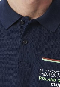 Marinblå polo skjorta med strukturerad tyg, två svarta knappar och en bröstficka med gröna, röda och ljusblå ränder samt broderad logotyp.