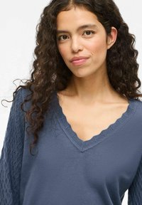 Suéter azul con escote en V con volantes y patrón texturizado en las mangas largas, hecho de una tela suave, que presenta una silueta ajustada.