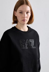 Μαύρη φόρμα με υφασματική λογότυπο "EA7 Emporio Armani", στρογγυλή λαιμόκοψη και χαλαρή εφαρμογή. Φτιαγμένη από μαλακό ύφασμα με ομαλή επιφάνεια.