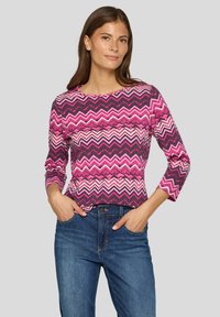 Langärmliges Oberteil mit rosa und lila Zickzack-Muster und Rundhalsausschnitt, kombiniert mit blauen Jeans aus Denim. Der Stoff scheint gerippt und hat einen figurbetonten Schnitt.