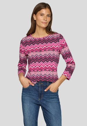 Langärmliges Oberteil mit rosa und lila Zickzack-Muster und Rundhalsausschnitt, kombiniert mit blauen Jeans aus Denim. Der Stoff scheint gerippt und hat einen figurbetonten Schnitt.