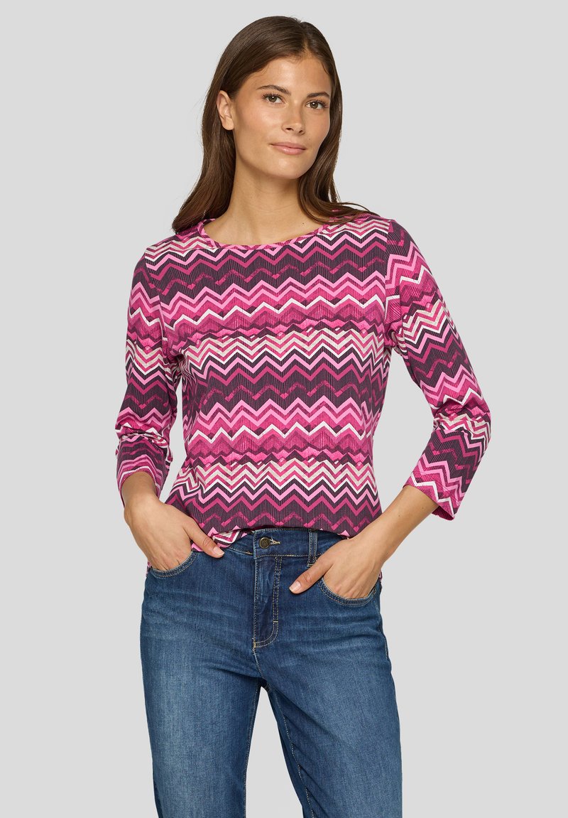 Langärmliges Oberteil mit rosa und lila Zickzack-Muster und Rundhalsausschnitt, kombiniert mit blauen Jeans aus Denim. Der Stoff scheint gerippt und hat einen figurbetonten Schnitt.