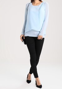 Light blue, long-sleeve top with pleats, layered design; paired with black skinny pants and black pointed-toe heels, holding a small black bag.