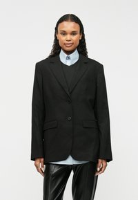 Čierny sakový blejzer s dvoma prednými gombíkmi, s výstrihom v tvare U, bočnými vreckami, nosený cez tmavú vestu a svetlomodrú košeľu s golierom. Hladká tkanina.