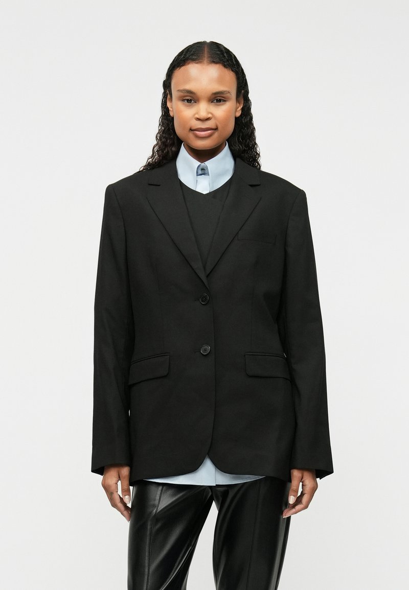 Čierny sakový blejzer s dvoma prednými gombíkmi, s výstrihom v tvare U, bočnými vreckami, nosený cez tmavú vestu a svetlomodrú košeľu s golierom. Hladká tkanina.