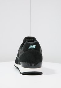 Sneaker nera New Balance con suola bianca, suola testurizzata e logo ricamato sul tallone, mostrata da dietro su una superficie bianca.