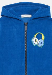 Zils korduroja zip-up jaka ar kapuci, melnu rāvējslēdzēju un karikatūristu donut grafiku dzeltenā, zilā un baltā krāsā uz krūtīm.