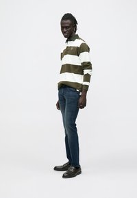 Długi rękaw koszulki polo w zielono-białe paski, granatowe jeansy, czarne mokasyny; model stoi na jednolitym tle, ręce w kieszeniach.