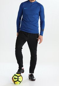 Maglia blu a maniche lunghe Nike in materiale performante con zip a metà, abbinata a pantaloni atletici neri e una palla da calcio giallo-verde brillante.