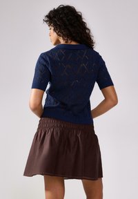 Maglione navy a maniche corte con motivo a rombi e bordi a coste, abbinato a una gonna marrone con vita arricciata. E' evidente un sottile contrasto di texture.
