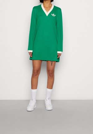 Osoba ubrana w zieloną sukienkę z długim rękawem i białym obszyciem, białe skarpetki i białe trampki, stojąca na jednolitym tle.