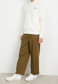 Felpa bianca con una toppa blu, abbinata a pantaloni beige a gamba larga e sneakers bianchi, con una texture liscia e un design casual.