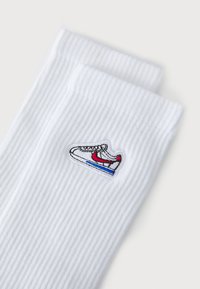 Fehér bordás zoknik, amelyek piros, fekete és kék hímzett sneaker mintával rendelkeznek. Simább textúrájúak és kényelmesen illeszkednek.