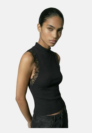 Žena s uhladenými vlasmi učesanými dozadu, oblečená v čiernom tielku bez rukávov s čipkovými bočnými panelmi a tmavých nohaviciach, stojaca pred jednoduchým pozadím.