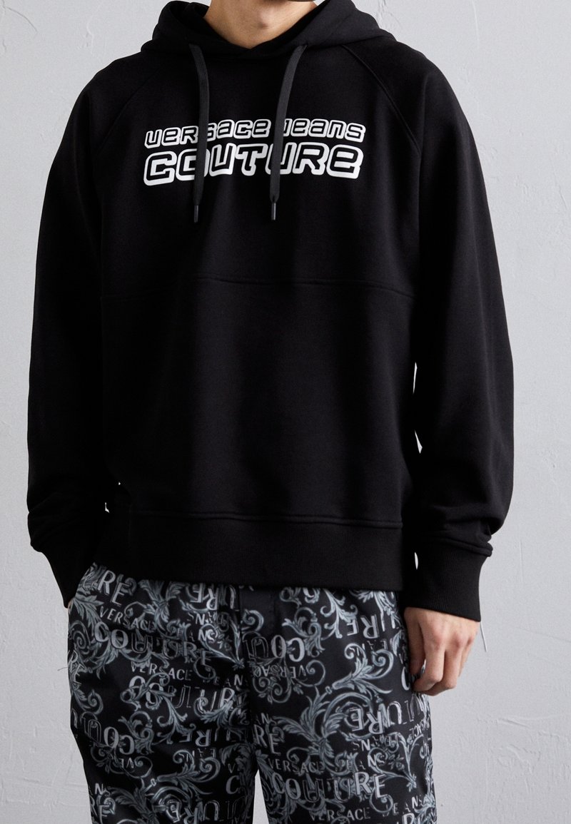 Černá mikina s kapucí z bavlněné směsi, s kapucí se šňůrkami a logem na přední straně. Kombinovaná s kalhotami s černo-šedým vzorem.