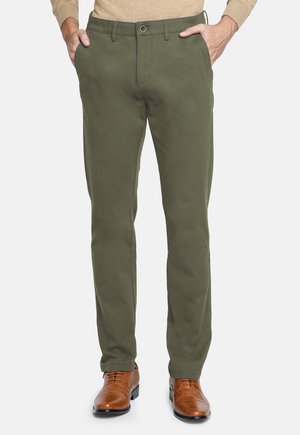 Hombre que lleva pantalones rectos de color verde oliva, suéter beige y zapatos de vestir de cuero marrón, con las manos en los bolsillos, de pie sobre un fondo blanco.