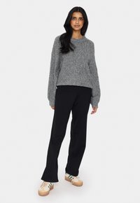 Grau gestrickter Pullover mit Rautenmuster, langen Ärmeln, kombiniert mit schwarzen Schlaghosen und beigen Plateausneakern mit grünen Streifen.