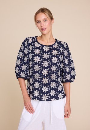 Kvinne iført en marineblå bluse med hvit blomsterbrodering og puffede trekvart-ermer, kombinert med hvite bukser.