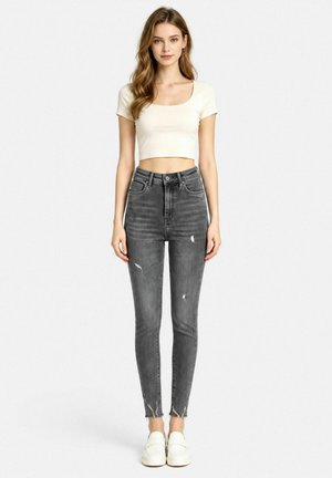 Giovane donna che indossa un top corto bianco, jeans skinny grigi strappati a vita alta e scarpe slip-on bianche, in piedi davanti a uno sfondo semplice.
