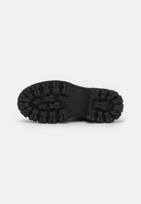 Suela de zapato de goma negra con un patrón de banda de rodadura profundo que presenta protuberancias rectangulares y circulares para tracción. Área central lisa sin patrón.