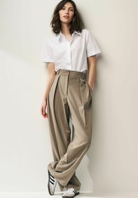 Chemise blanche légère à boutons, associée à un pantalon marron à rayures. Le modèle porte des baskets noir et blanc, posant contre un mur.