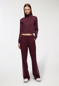 Bordowy dwuelementowy dres, w skład którego wchodzi krótkia kurtka z zamkiem błyskawicznym i wysokim kołnierzem oraz dopasowane spodnie z białymi paskami po bokach. Gładka tekstura.