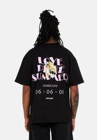 Camiseta negra de gran tamaño con gráficos impresos coloridos en la parte posterior, que incluyen el texto "AMOR VERANO" y "VERANO AMOR", acompañados de elementos de dibujo animado.