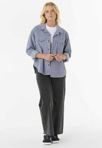 Giacca azzurra chiara con bottoni, due tasche sul petto, vestibilità comoda e maniche arrotolate. Abbinata a pantaloni grigio scuro e scarpe da ginnastica nere.